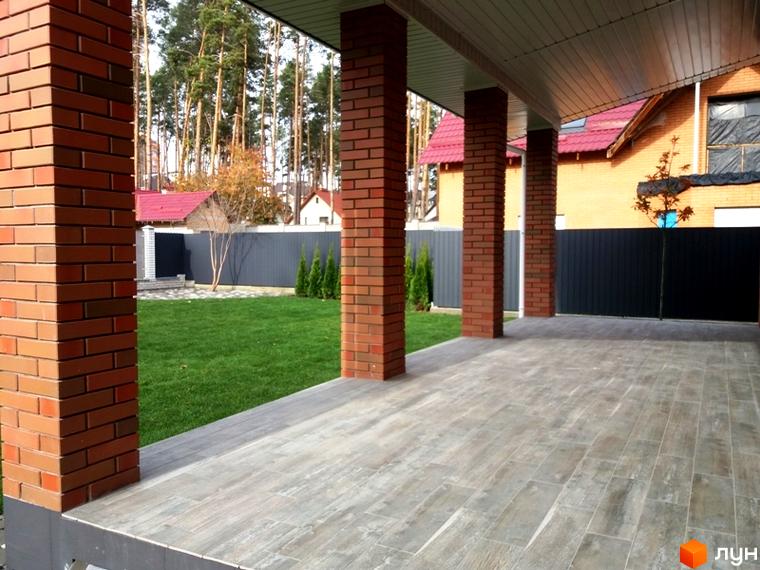 Зовнішній вигляд будинку з цегляними колонами та терасою, що виходить на зелену прибудинкову територію з деревами та парканом.