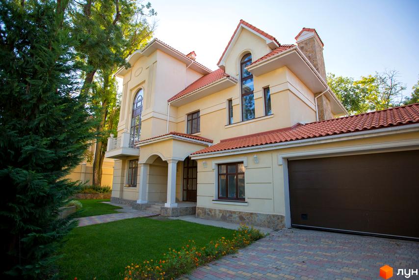 Двоповерховий будинок з червоною черепицею та гаражем. Перед будинком газон та доріжка з бруківки, навколо зелені дерева.