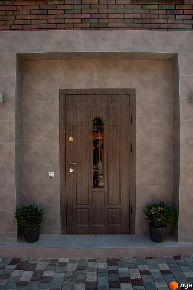 Таунхаус „На Кочубея“: вхідна група з дерев'яними дверима та вікном, вимощена доріжка та зелені кущі. Новобудова в передмісті Києва.