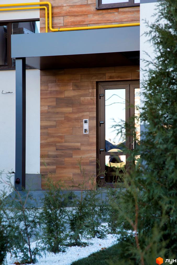 Вхідна група в будинок з дерев'яним оздобленням стін та скляними дверима. На прибудинковій території висаджені кущі та білий камінь.