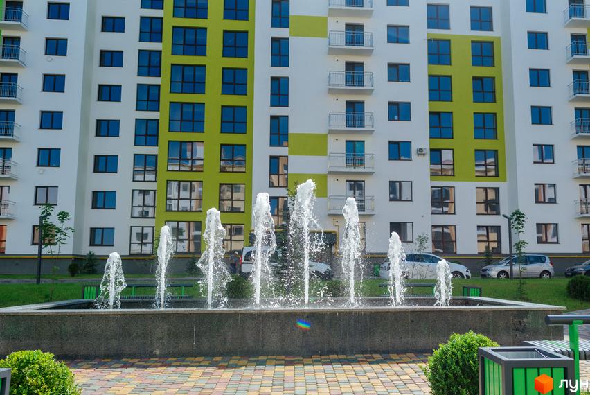 Багатоповерховий будинок Житлового комплексу На Щасливому з білим та салатовим фасадом, великими вікнами та балконами. Перед ним фонтан та кольорова бруківка.