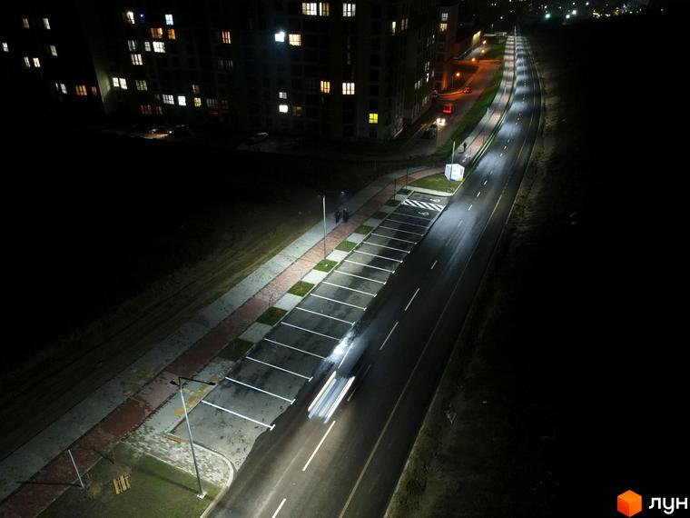 Багатоповерховий будинок Житлового комплексу На Щасливому вночі з освітленими вікнами. Поруч освітлена дорога, парковка та тротуар.