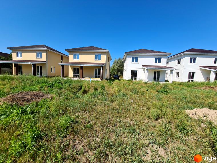 Котеджне містечко КМ Garden House: двоповерхові будинки з коричневими дахами та білими стінами на тлі блакитного неба. Навколо будинків зелена трава та кущі.