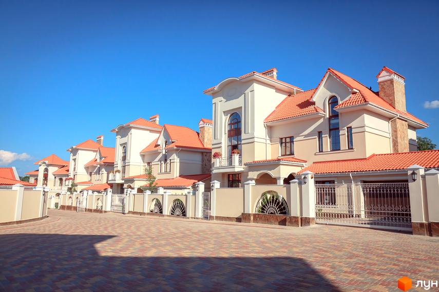 Котеджне містечко Oasis з будинками з червоною черепицею та світлими фасадами. Територія вимощена бруківкою, огороджена білим парканом з кованими елементами.