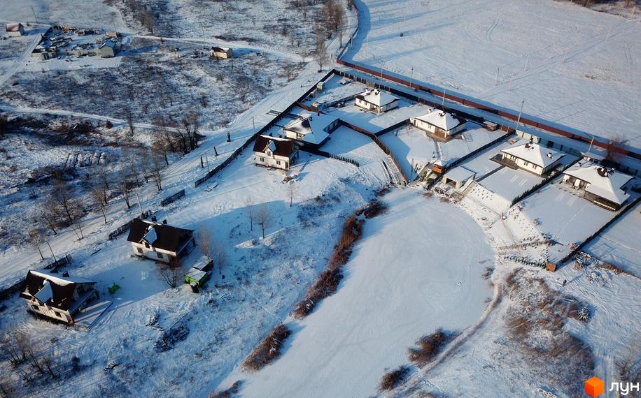 Котеджне містечко КМ Маєток взимку. Білі будинки з темними дахами розташовані на засніженій ділянці біля замерзлого озера. Навколо будинків паркани та дерева.