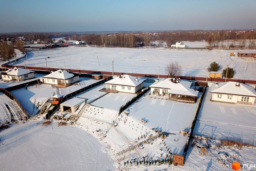 Котеджне містечко КМ Маєток. Білі одноповерхові будинки з коричневими дахами, вкриті снігом. Навколо будинків доглянуті ділянки з деревами та доріжками.