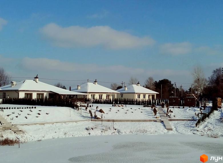 Котеджне містечко КМ Маєток. Три одноповерхові будинки з білими стінами та темними дахами, вкритими снігом, розташовані на схилі пагорба. Навколо будівель висаджені туї, а перед ними замерзле озеро.