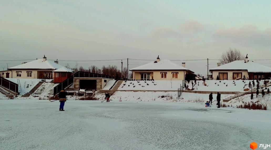 Котеджне містечко КМ Маєток: одноповерхові будинки з білими стінами та сірими дахами, вкритими снігом. Навколо будівель – засніжена територія з деревами та людьми, що відпочивають.