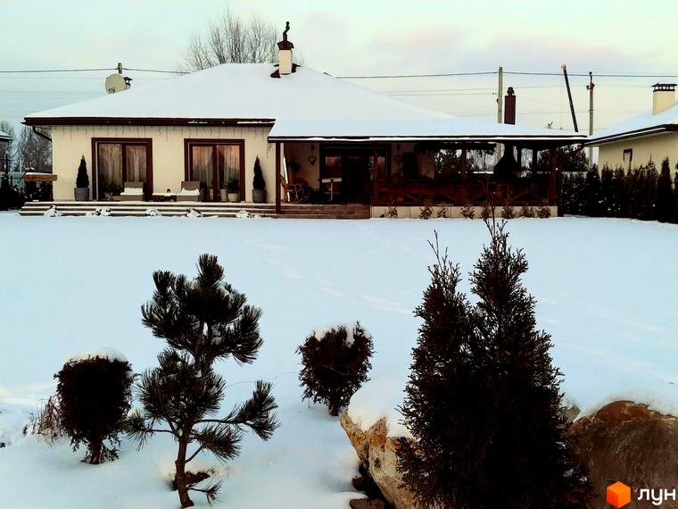 Котеджне містечко КМ Маєток взимку. Одноповерховий будинок з великою терасою, вкритий снігом, на фоні засніженої ділянки з ялинками та сосною.