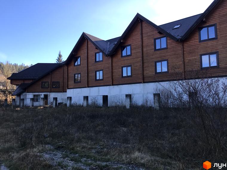 Котеджне містечко КМ Семидвір. Двоповерховий дерев'яний будинок з великими вікнами на тлі блакитного неба. Навколо будинку - зарості трави та кущів.