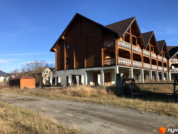 Котеджне містечко КМ Семидвір. Будівля з дерев'яних брусів з балконами та коричневим дахом на тлі блакитного неба. Навколо будівлі суха трава та ґрунтова дорога.