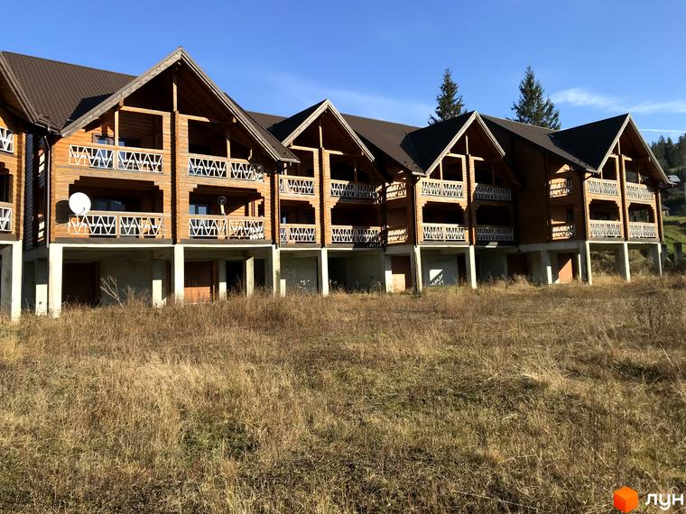 Котеджне містечко КМ Семидвір: дерев'яні будинки з балконами на тлі блакитного неба та зелених пагорбів. Навколо - суха трава та дерева.