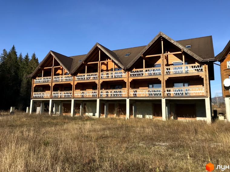 Котеджне містечко КМ Семидвір – триповерховий дерев'яний будинок з балконами та терасами на тлі лісу та блакитного неба. Навколо – висока суха трава.