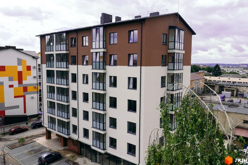 Сучасний житловий комплекс на вулиці Транспортна, 10, з біло-коричневим фасадом та великими вікнами, поруч парковка та сусідня будівля з яскравим геометричним візерунком.