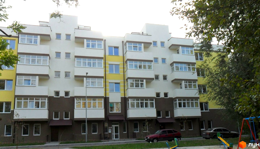 Багатоповерховий будинок з білими балконами та вікнами. На території є зелена зона та дитячий майданчик.