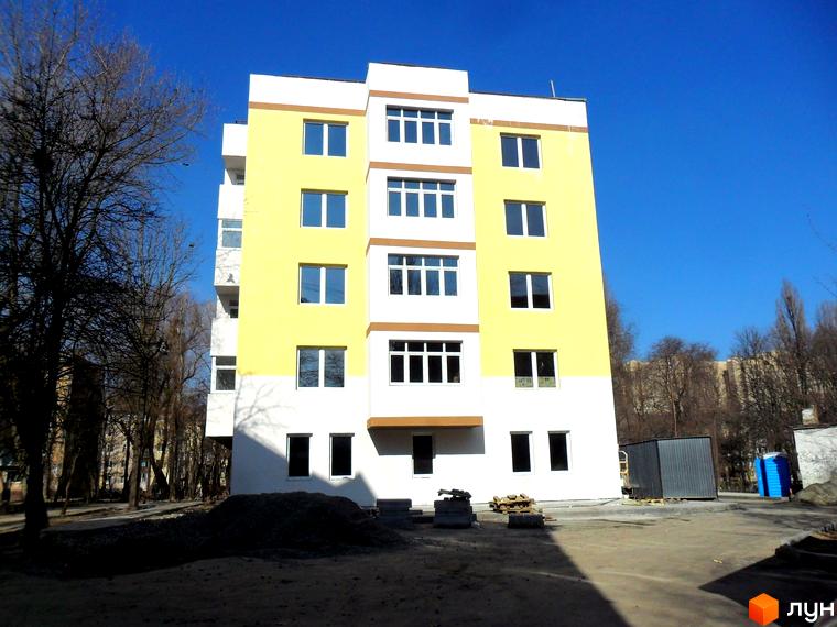 Багатоповерховий будинок з жовтим та білим фасадом на тлі блакитного неба. На передньому плані видно будівельні матеріали та дерева.