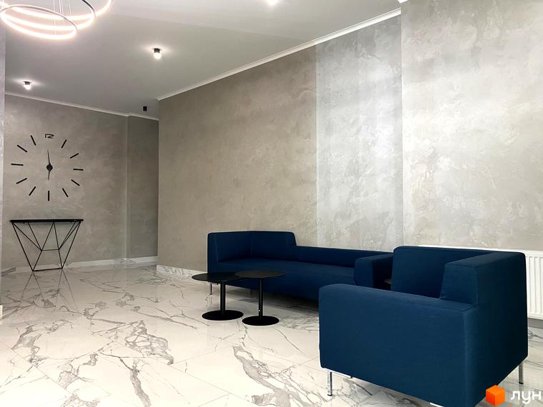 Дизайн холу житлового комплексу Корфу з синіми диванами та круглими столиками. На стіні великий настінний годинник.