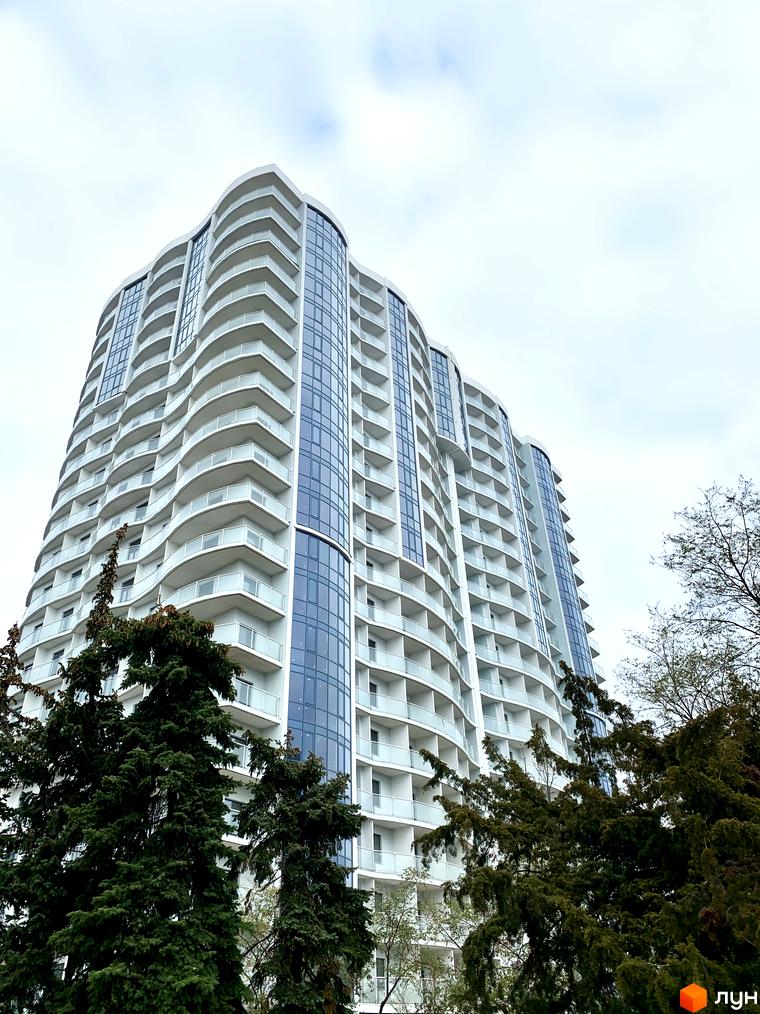 Зображення багатоповерхового будинку Житлового комплексу Корфу з балконами та панорамними вікнами. Навколо будинку ростуть дерева.