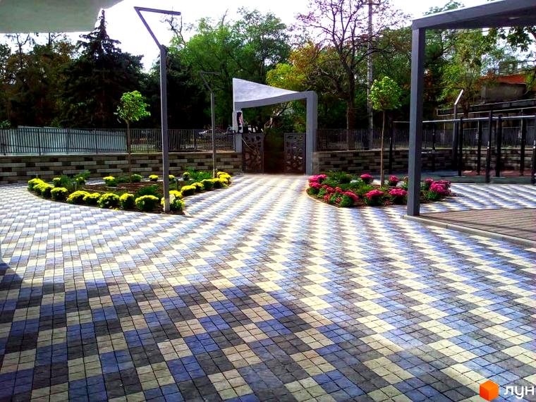 Зображення території Житлового комплексу Корфу з тротуарною плиткою, клумбами з квітами та декоративними деревами. Вигляд на вхідну групу та елементи благоустрою.