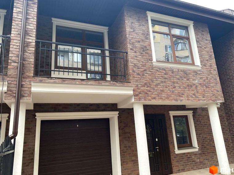 Зовнішній вигляд таунхауса в ЖК „Протасів Residence“ з цегляним фасадом, балконом та гаражем. Простір навколо будинку з доглянутою прибудинковою територією.