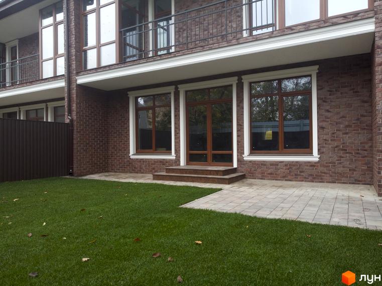 Цегляний будинок з великими вікнами та балконом. Перед будинком газон та плитка, що веде до входу.