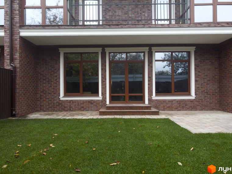 Фасад будинку з коричневою цегляною кладкою та великими вікнами. Перед будинком газон з травою та бруківка.