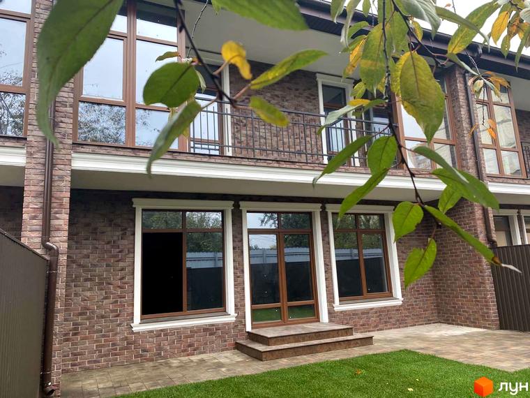 Зовнішній вигляд таунхаусів Протасів Residence з цегляною кладкою, великими вікнами та балконом. На передньому плані - зелена трава та гілки дерев.