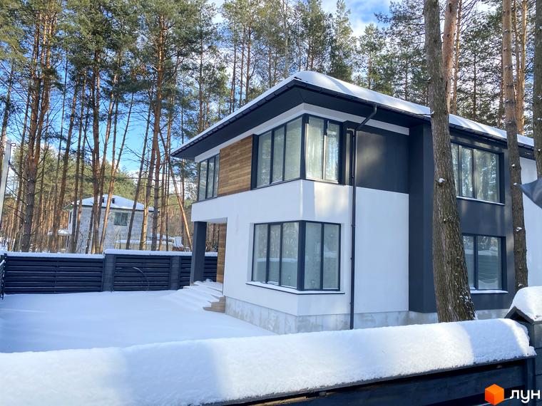 Двоповерховий будинок з білими стінами та темною покрівлею в Котеджному містечку Квітка Полонини, вкритий снігом. Навкруги будинку ростуть дерева.
