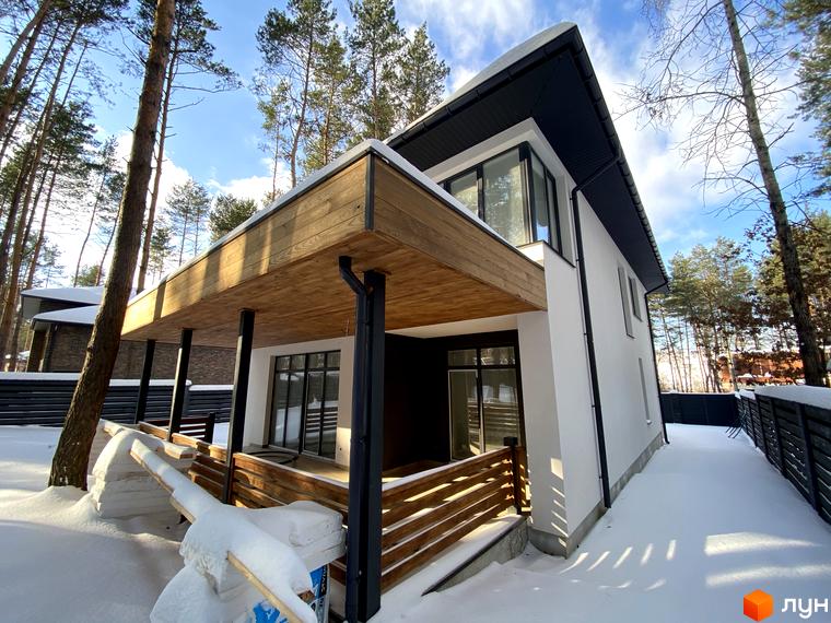Котеджне містечко Квітка Полонини. Білий двоповерховий будинок з дерев'яною терасою та огорожею, вкритий снігом. Навкруги ростуть дерева.