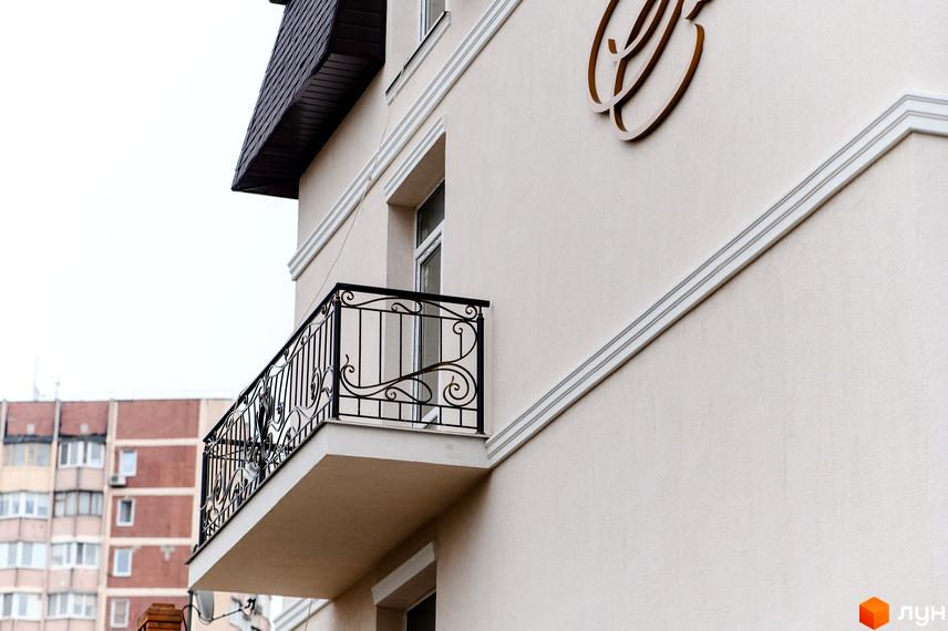 Фасад будинку з балконом та кованими перилами. На стіні будівлі розташований логотип. Поруч видно вікна та інші елементи архітектури.
