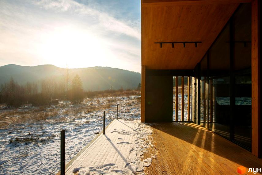Дерев'яний будинок з панорамним склінням та терасою, вкритий снігом. На тлі зимовий пейзаж з горами та сонячним світлом. КМ INO club.