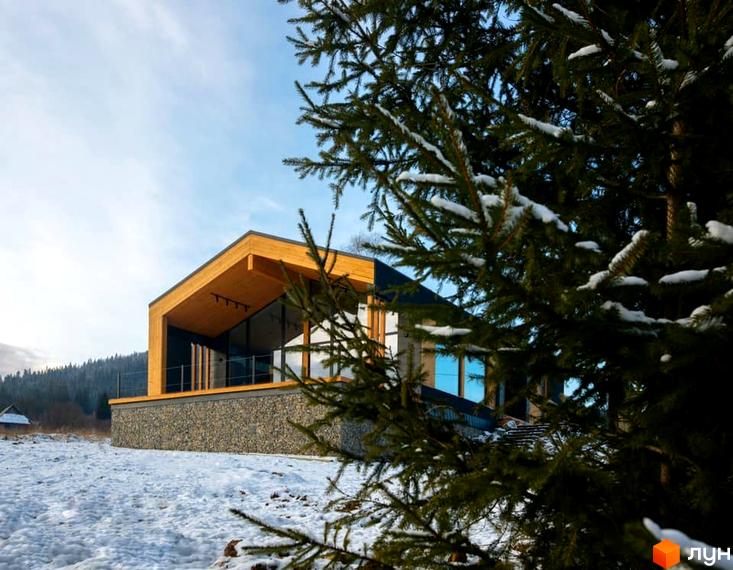 Будинок з дерева та скла в Котеджному комплексі INO club, розташований серед засніженої місцевості. Навколо будівлі видно хвойні дерева та сніг.