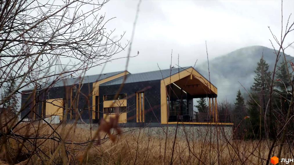 Зображення будинку КМ INO club з темним фасадом та дерев'яними елементами. Будинок розташований серед гірської місцевості, на передньому плані видно гілки дерев.