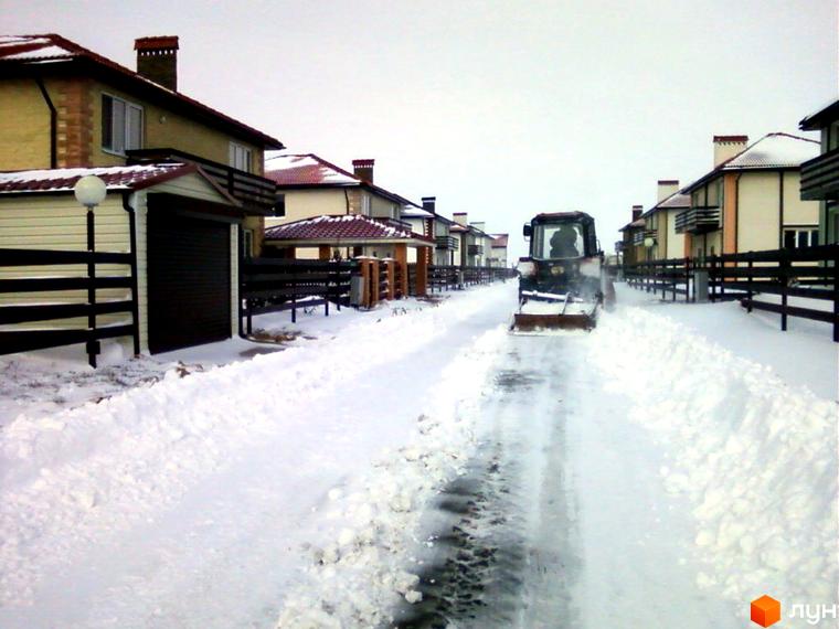 Зимовий пейзаж житлового комплексу Fresh Town. Дорога очищається від снігу трактором, навколо будинки з червоними дахами.
