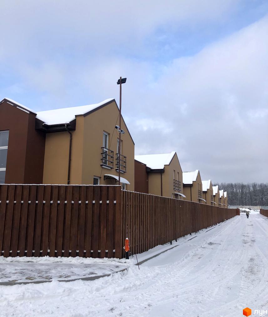 Котеджне містечко Sunny Village: фасади будинків коричневі та бежеві, дахи вкриті снігом. Перед ними дерев'яний паркан та засніжена дорога.