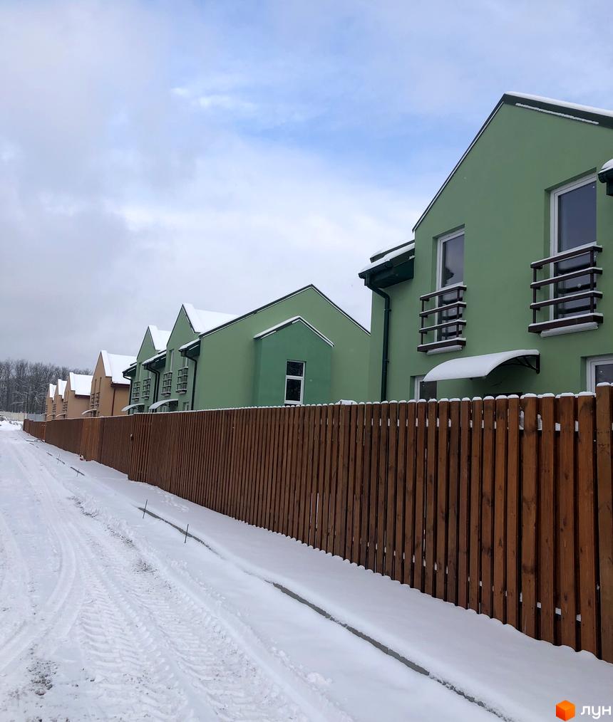 Зелені та бежеві будинки Котеджного містечка Sunny Village із засніженими дахами та дерев'яним парканом уздовж сніжної дороги.