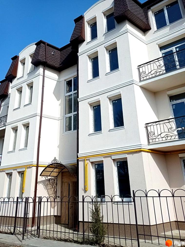 Білосніжний будинок Житлового комплексу з темним дахом та кованими балконами. Перед будинком кований паркан та зелені насадження.