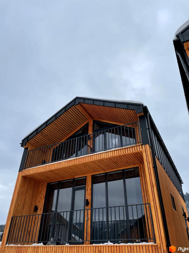 Дерев'яний будинок Котеджного містечка Phoenix Relax Park з темним дахом, великими вікнами та балконами з чорними перилами. Навколо видно сніг.