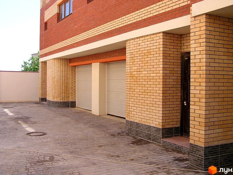 Таунхаус „Морський“ з фасадом з червоної та світлої цегли, темним цоколем, двома гаражними воротами та чорними вхідними дверима. Територія викладена бруківкою.