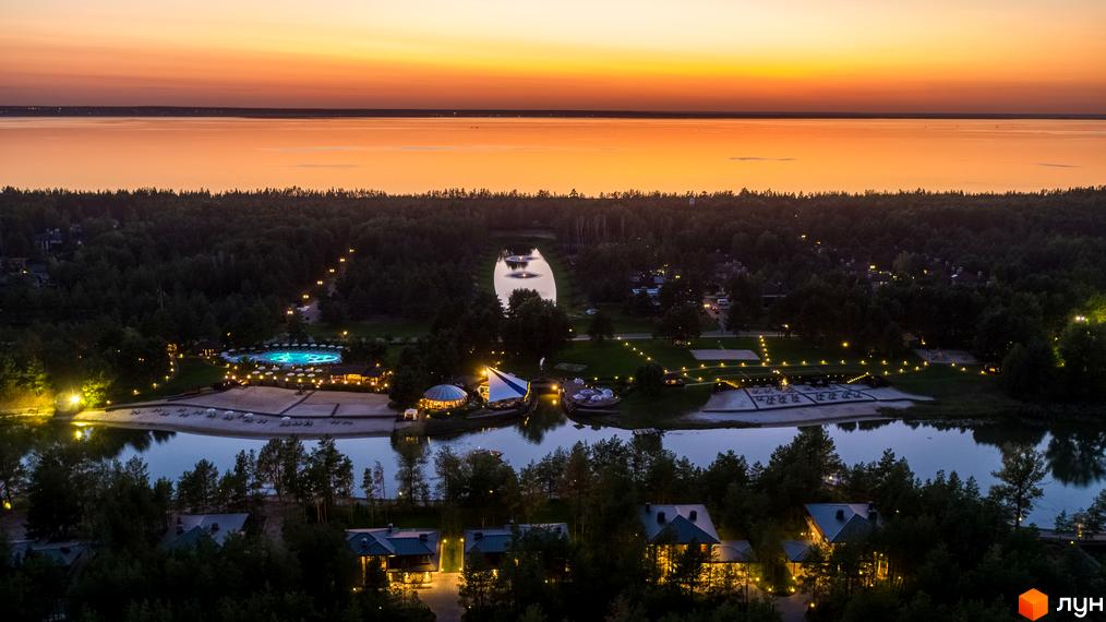 Вид на житловий комплекс КМ Riviera Village з вечірнім освітленням, прибудинкова територія з водоймою та зеленими насадженнями.
