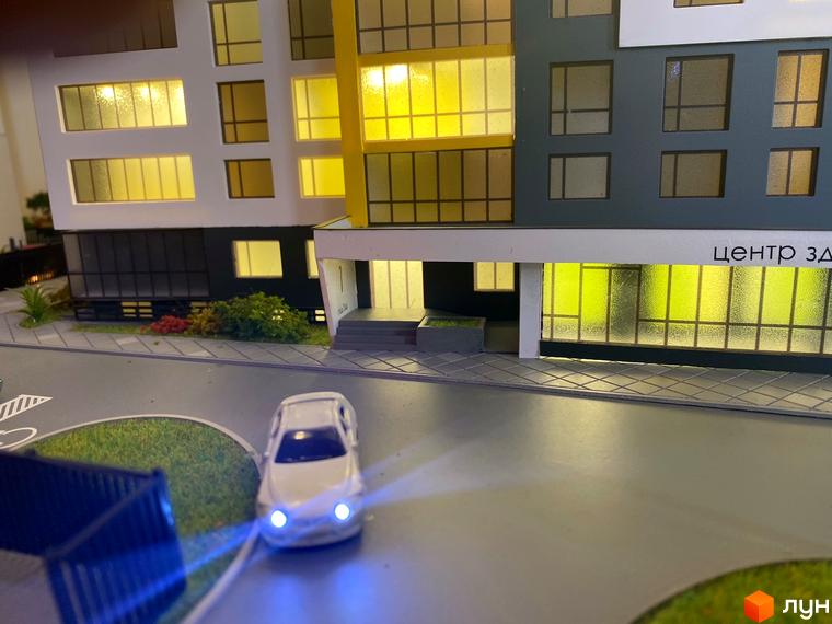 Зображення сторінки ЖК Квартал Енергія. На фото модель будинку з великими вікнами та освітленням, перед будинком дорога та автомобіль.