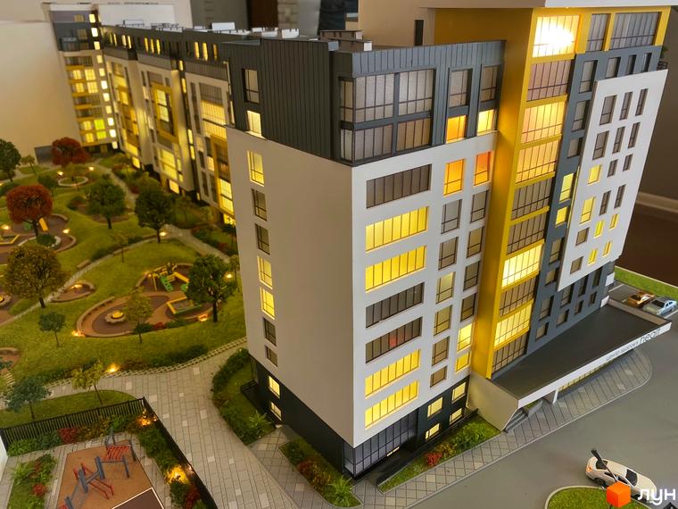 Макет багатоповерхового будинку з великими вікнами та жовтими вставками. Поруч розташована зелена прибудинкова територія з деревами, дитячим майданчиком та доріжками.