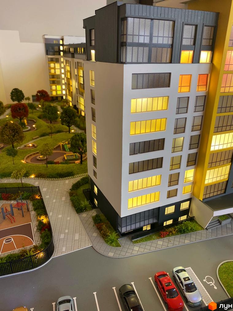 Макет житлового комплексу з освітленими вікнами, поруч дитячий майданчик, алеї для прогулянок, парковка з автомобілями.
