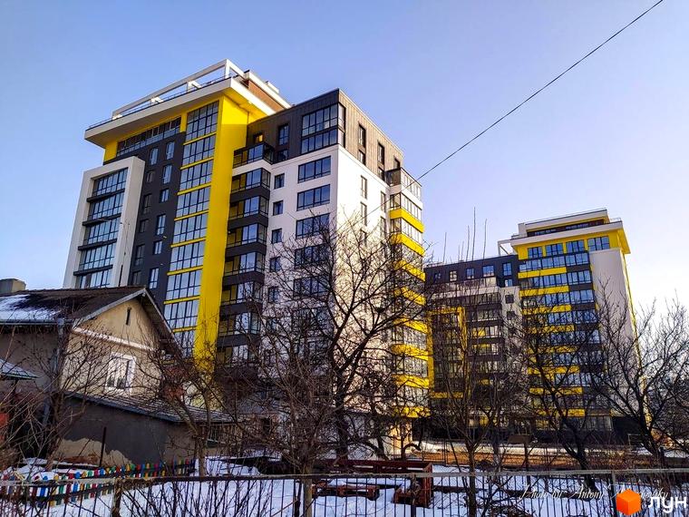 Житловий комплекс Енергія - це сучасна новобудова з яскравим жовтим фасадом та великою кількістю вікон. Навколо комплексу розташовані дерева та паркан.