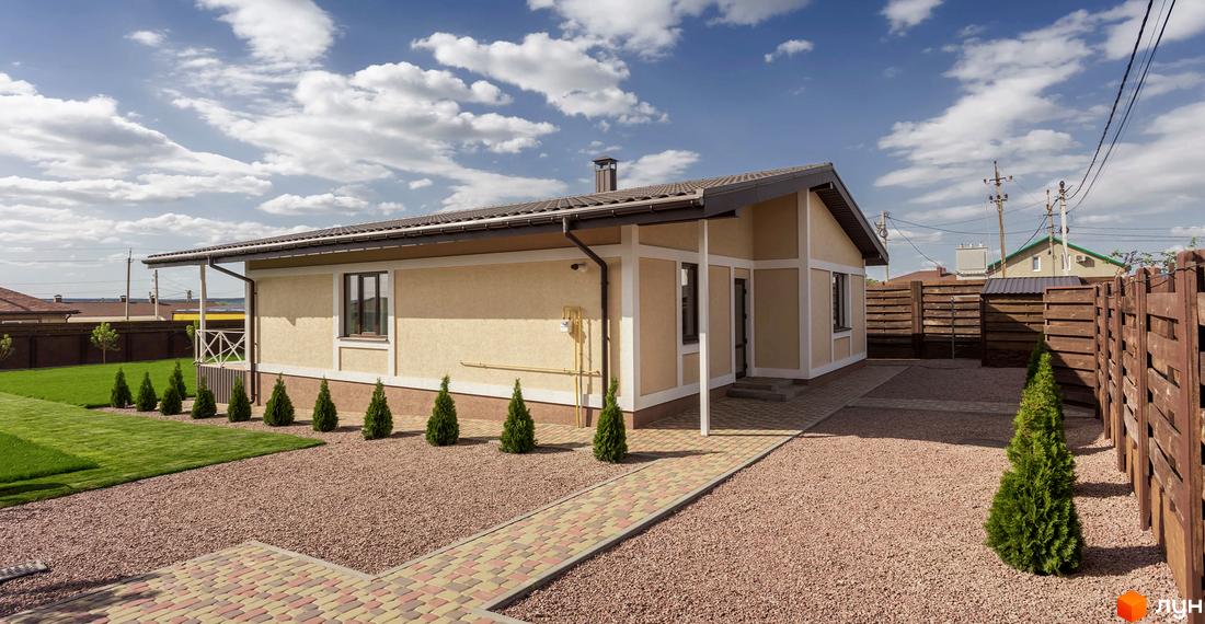 Одноповерховий будинок світло-бежевого кольору з коричневим дахом та вікнами в Котеджному містечку Графський. Перед будинком доріжка з бруківки, гравійне покриття та зелений газон.