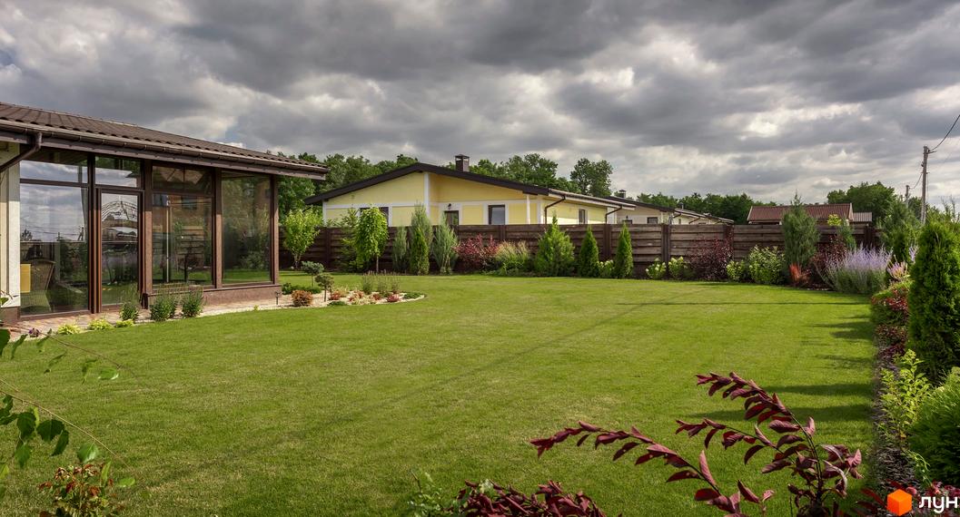 Будинок Котеджного містечка Графський з великими скляними вікнами та коричневим дахом. Навколо доглянутий зелений газон та різноманітні рослини.