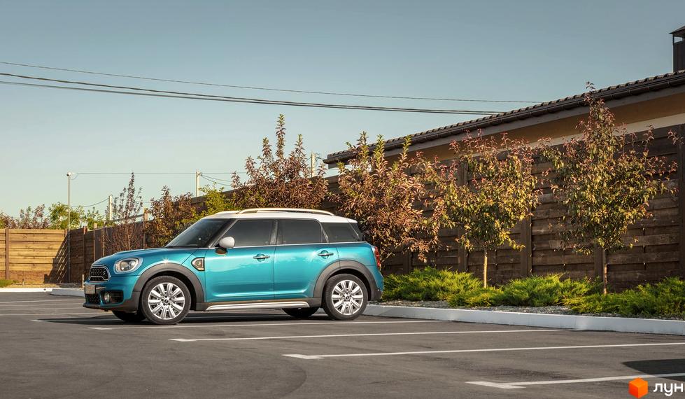 Бірюзовий автомобіль Mini Cooper Countryman на парковці біля дерев'яного паркану та зелених насаджень. Вид збоку на авто.