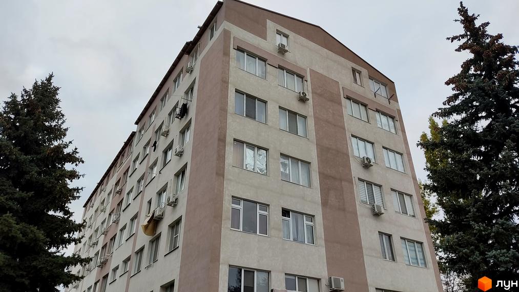 Багатоповерховий будинок ЖК Будинок на Боровського світлого кольору з коричневими вставками. Поруч із будівлею ростуть високі хвойні дерева.