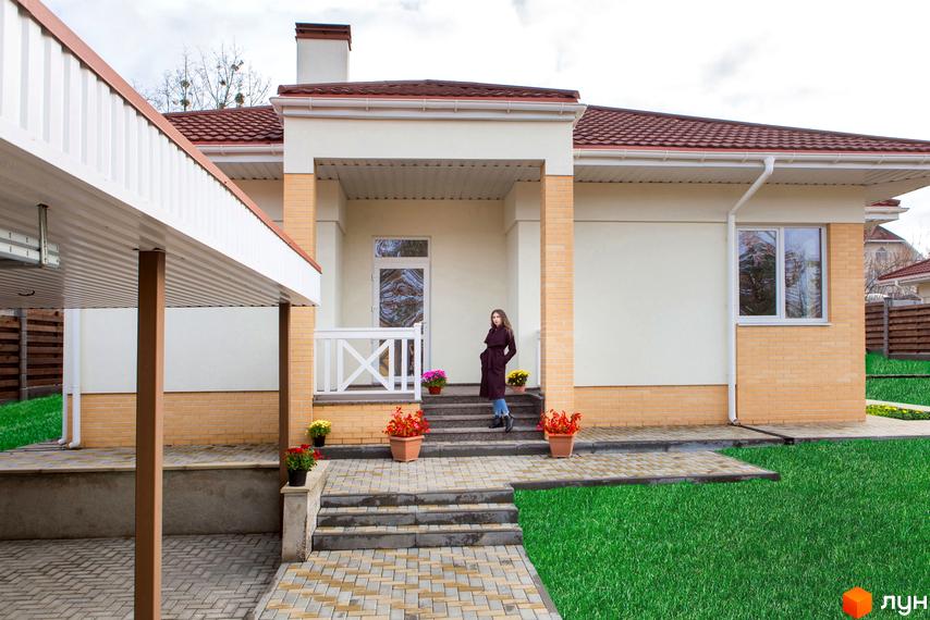 Вигляд будинку в Котеджному містечку Country House з газоном та бруківкою. На сходах до будинку стоять вазони з квітами.