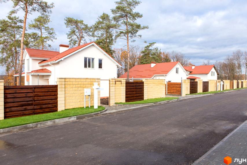 Котеджне містечко Country House: білі будинки з червоними дахами, паркани з цегли та дерева, зелений газон і асфальтована дорога.